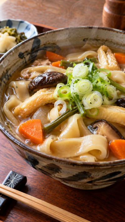 東京都奥多摩地域の郷土料理のしこみうどんを幅広の手打ち麺とねぎ、人参、しいたけ、油揚げなどで具だくさんに仕上げ、和食器に盛り付けた料理